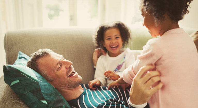 Man lays on the sofa with playing with twin daughters.